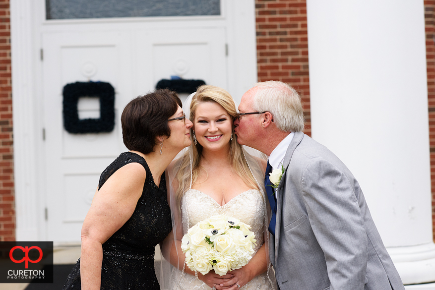 Bride's parents kiss her on the cheek.