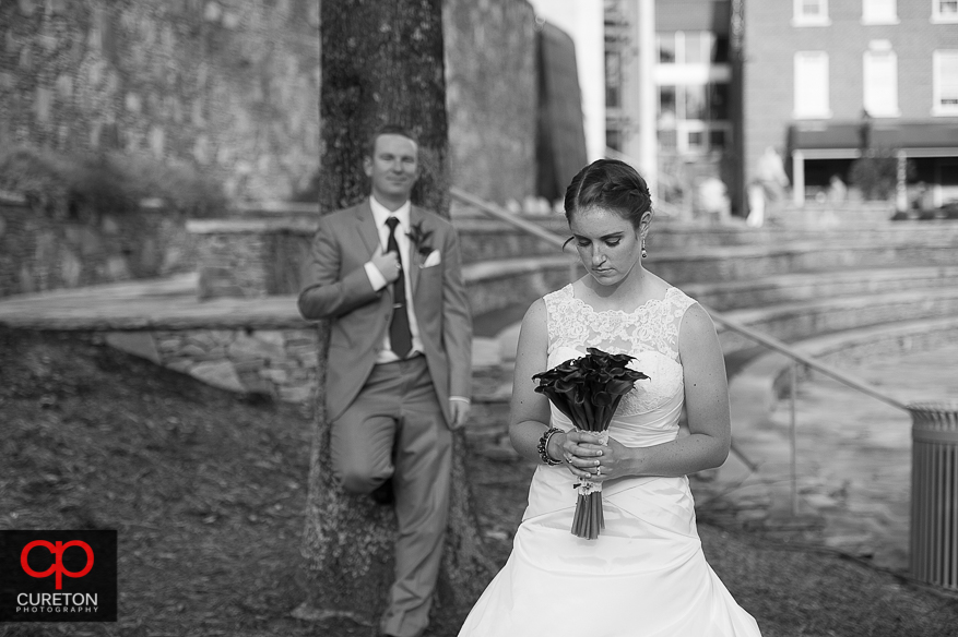 The bride looks at her flowers while the groom stands in the distance.