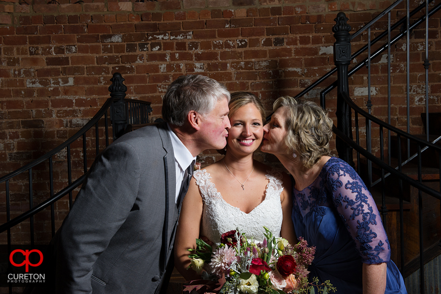 The brides parents kissing her on the cheek.