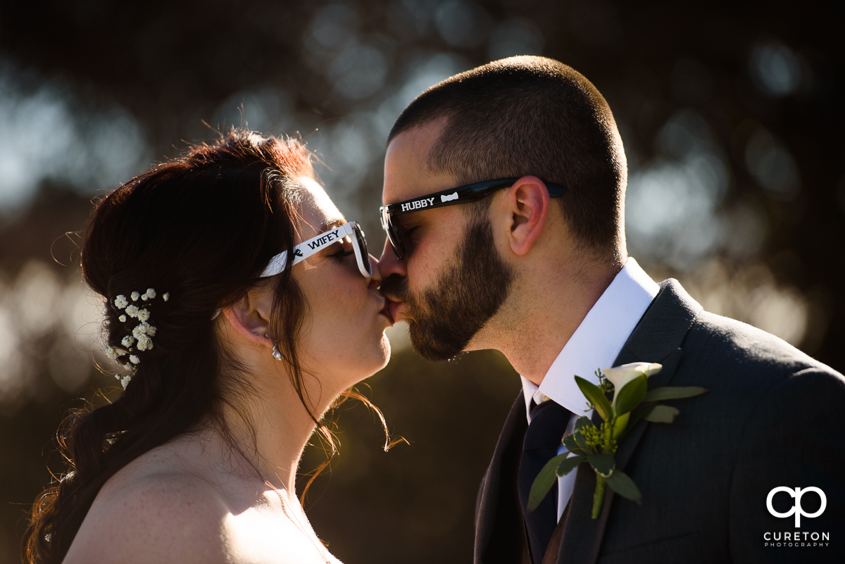 Bride and groom kissing while wearing printed sunglasses with the words wifey and hubby on them.