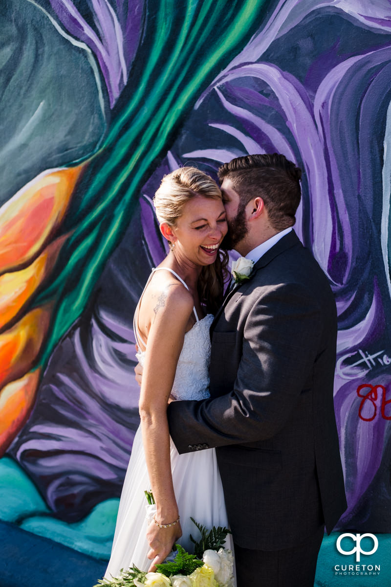 Bride laughing while her groom whispers in her ear outside Artisan Traders in Greenville.