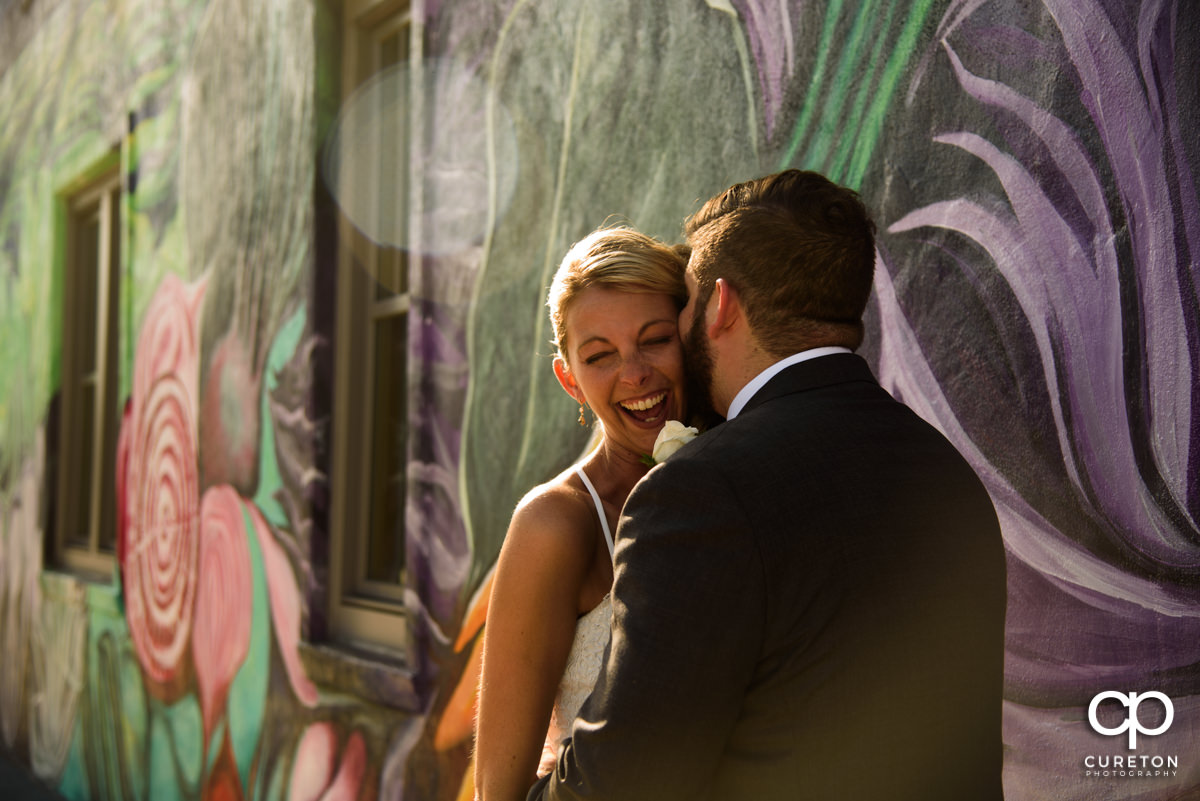 Groom kissing his bride on the cheek making her laugh while standing in front of a mural in downtown Greenville,SC.