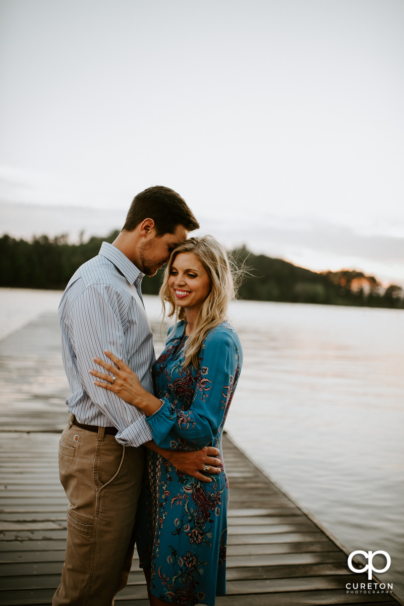 Man holding his fiancee on a dock by the lake at sunset at their engagement session in Clemson.