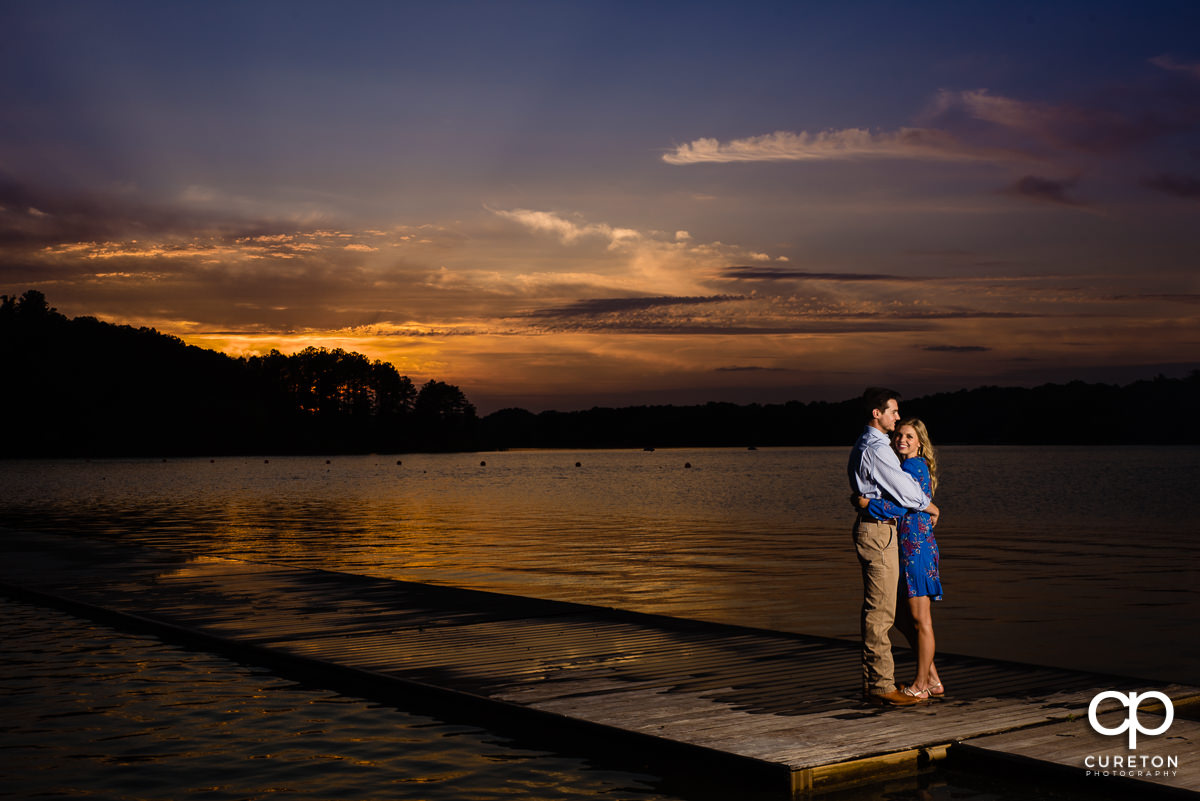Engaged couple hugging on a dock by the lake on the Clemson University campus at sunset.