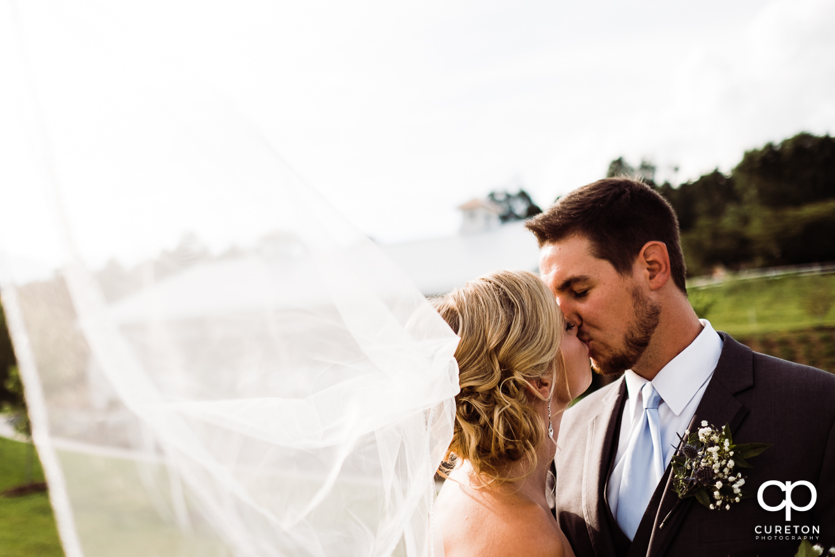 Groom kissing his bride with her veil blowing in the wind.