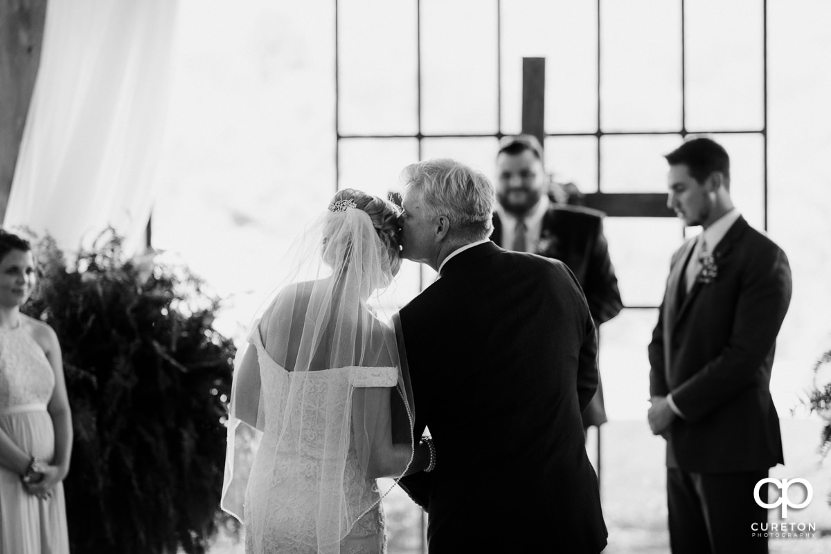 Bride's father kissing her on the forehead as he gives her away at the ceremony.