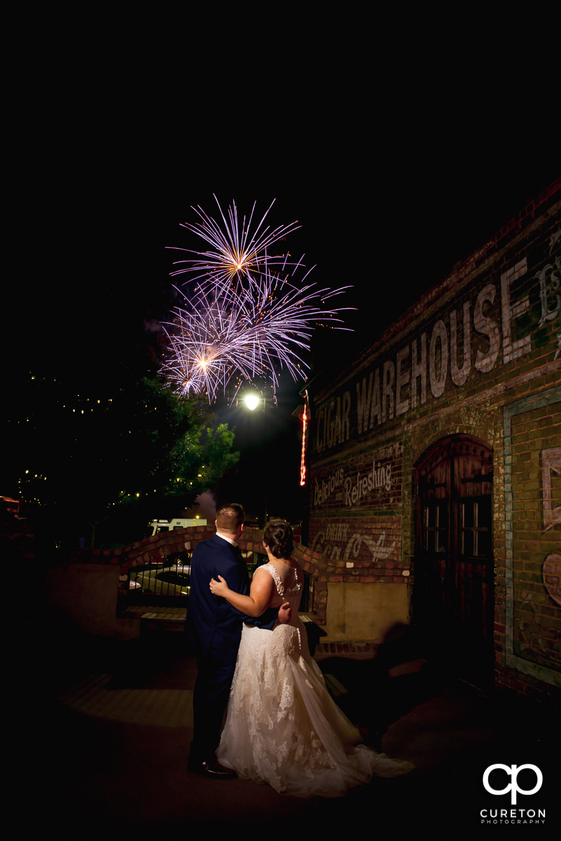 Bride and groom watching the fireworks show from the deck of The Old Cigar Warehouse after their wedding in downtown Greenville .