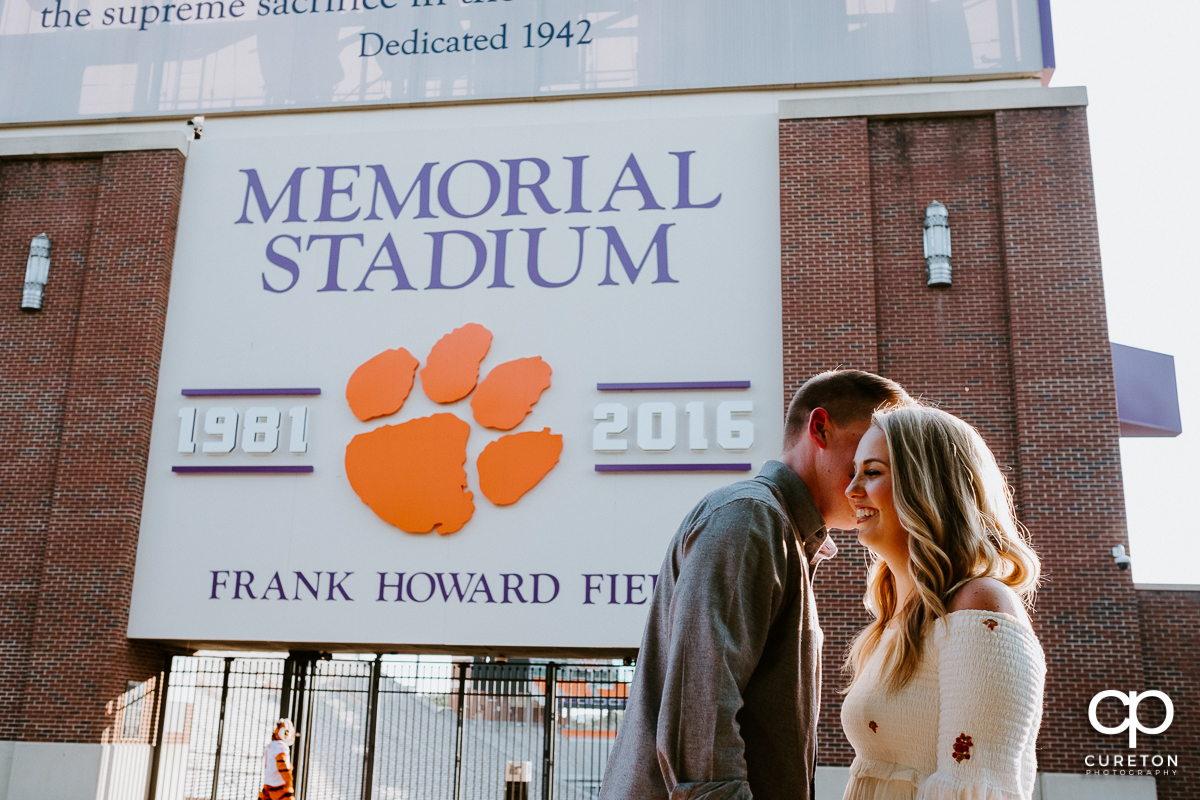 Man making his future bride laugh in front of a football stadium.