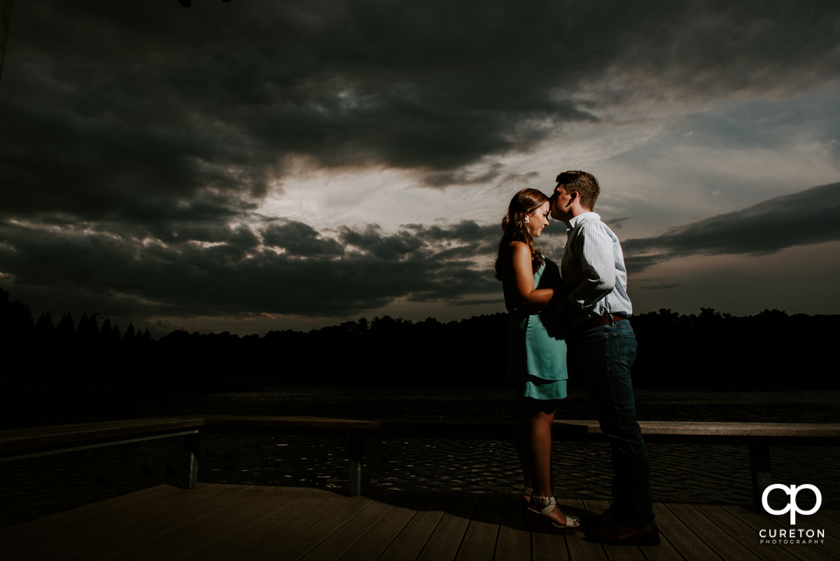 Future groom kissing his bride on the forehead at sunset by the lake during their creative engagement session.