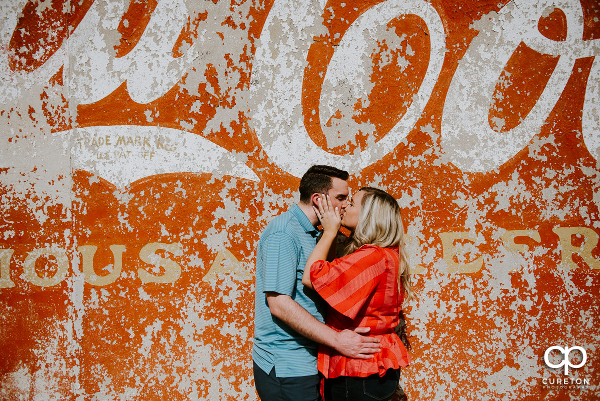Engaged couple kissing beside a vintage coca cola advertisement on a wall.