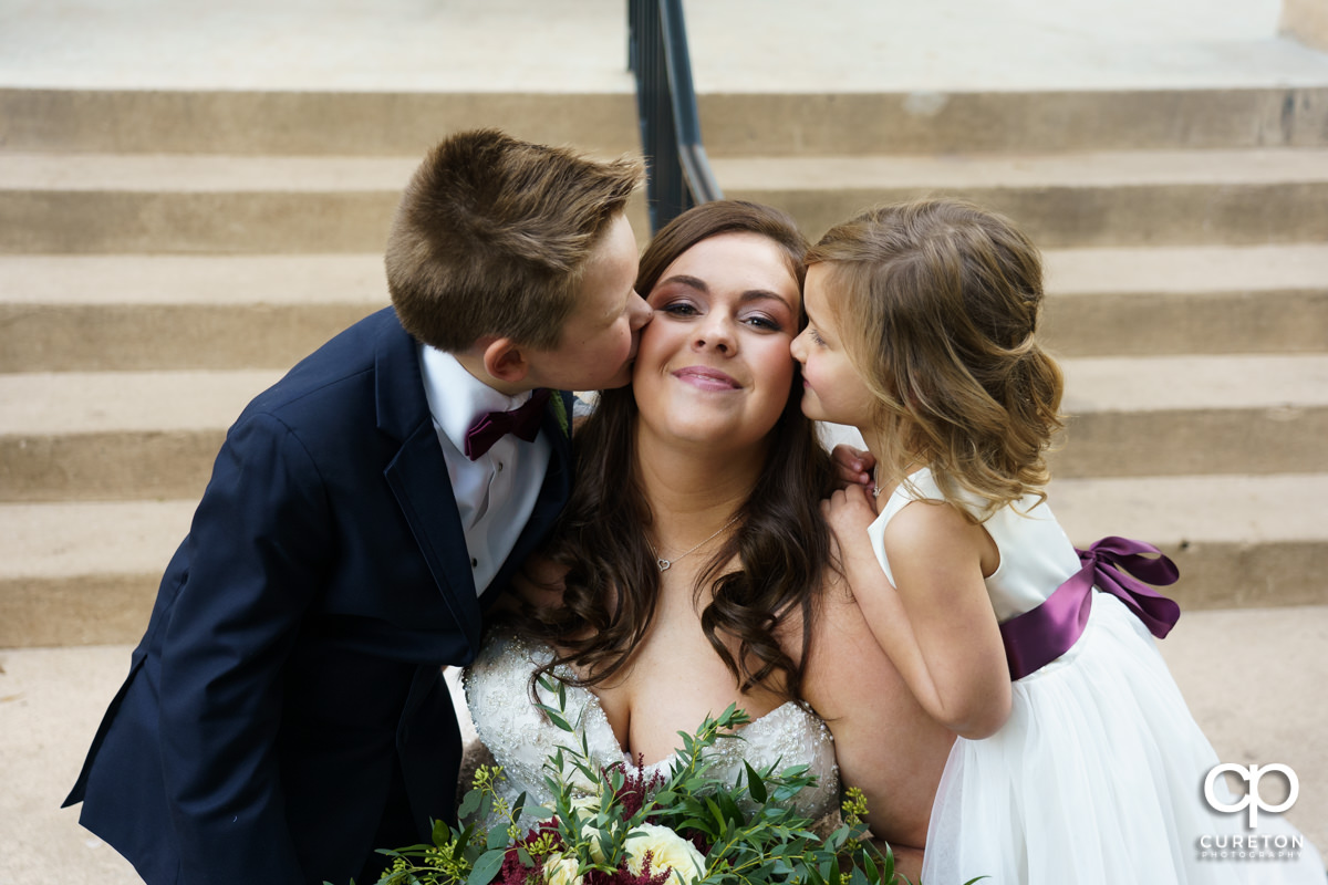Flower girl and ring bearer kissing the bride on the cheek.