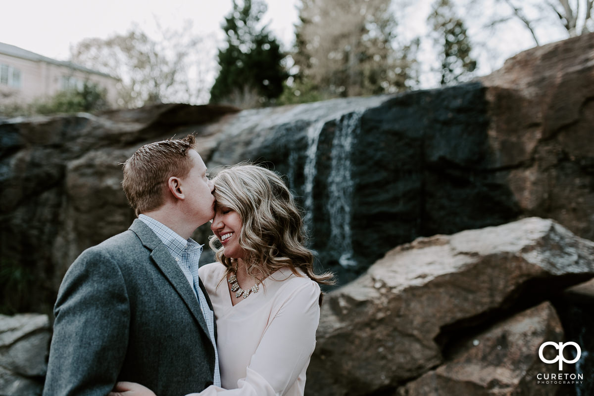 Future groom kissing his bride on the forehead in front of a waterfall during their engagement session.