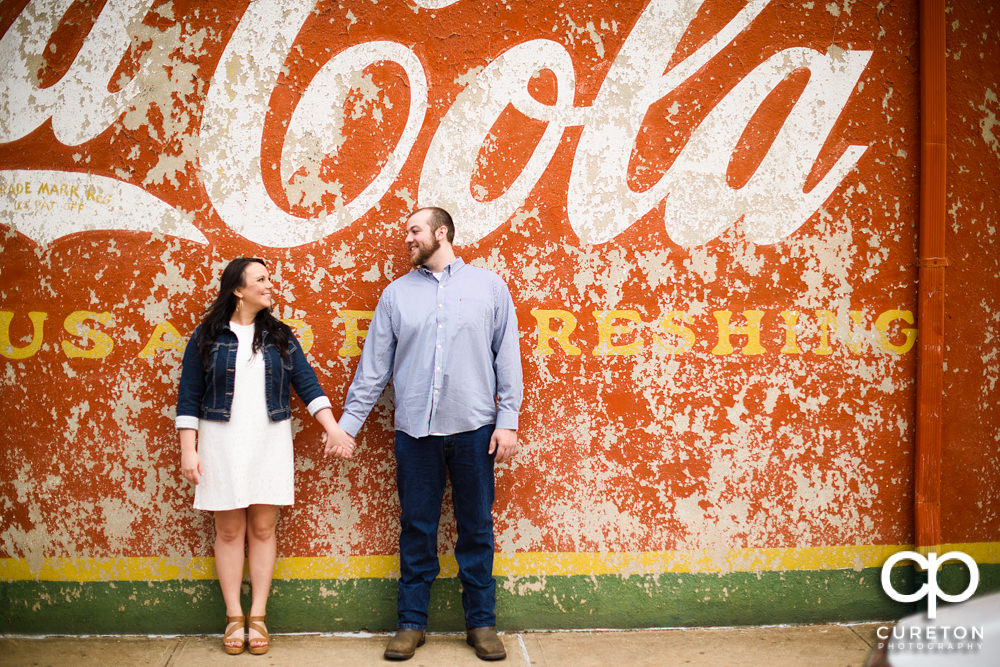 Engaged couple holding hands in front of the Coca Cola wall in downtown Greenville,SC.
