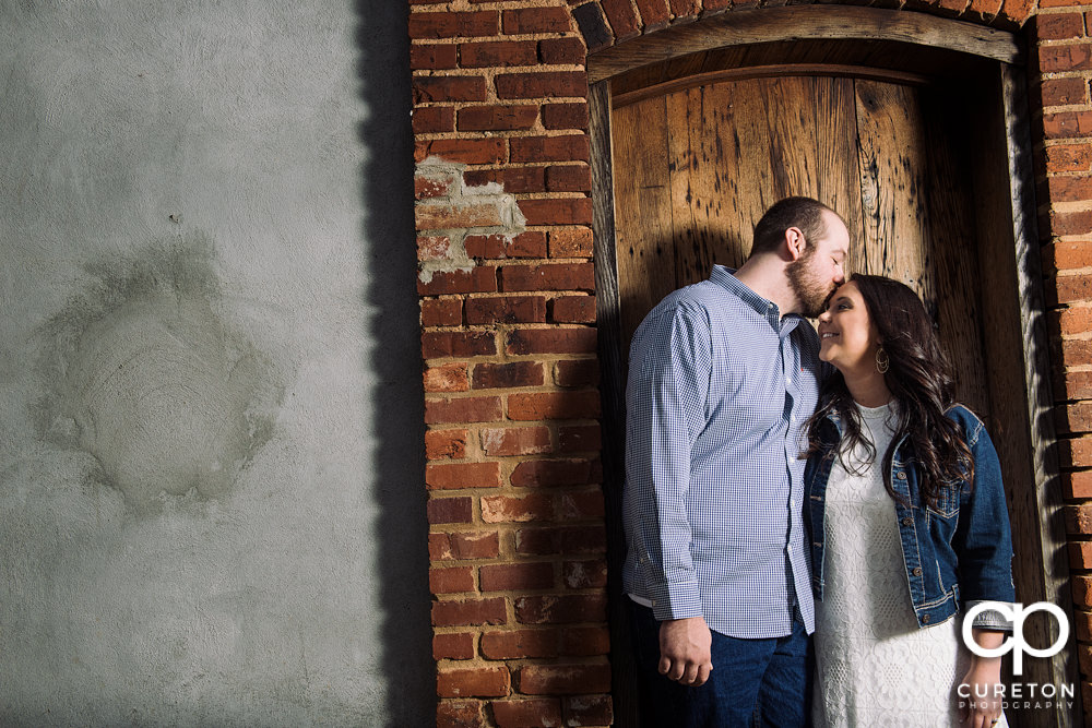 Man kissing his fiancee on the forehead inside a wooden doorway.