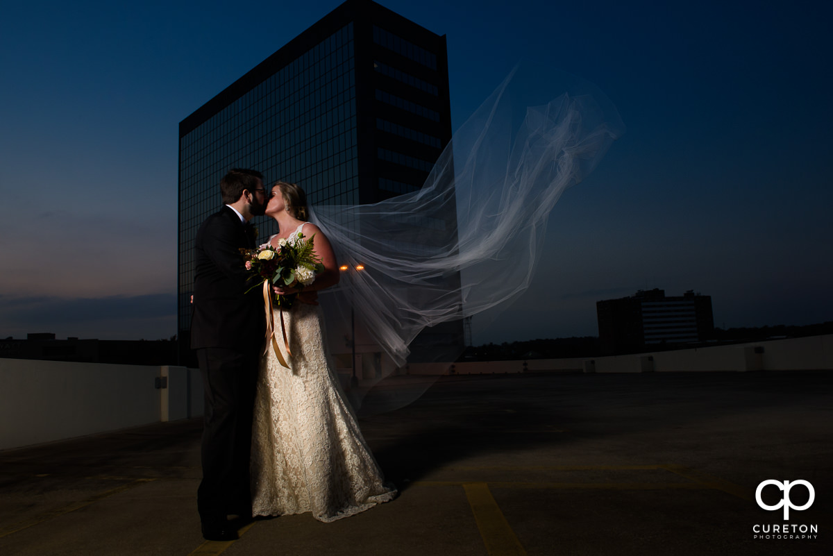 Bride and groom kissing on a rooftop with the veil blowing in the wind after their wedding at The Commerce Club in downtown Greenville, SC.