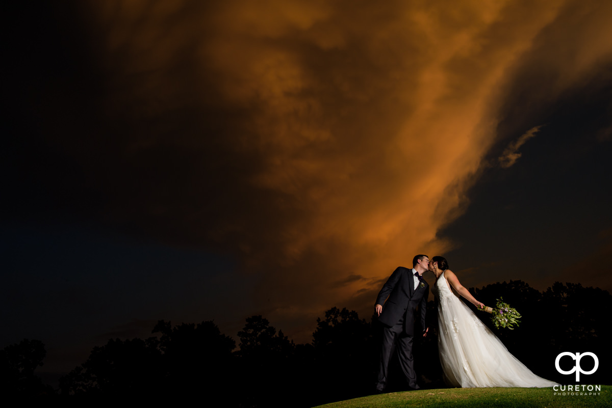 Bride kissing her groom at sunset under an amazing sky after tier wedding ceremony at Holly Tree in Simpsonville,SC.
