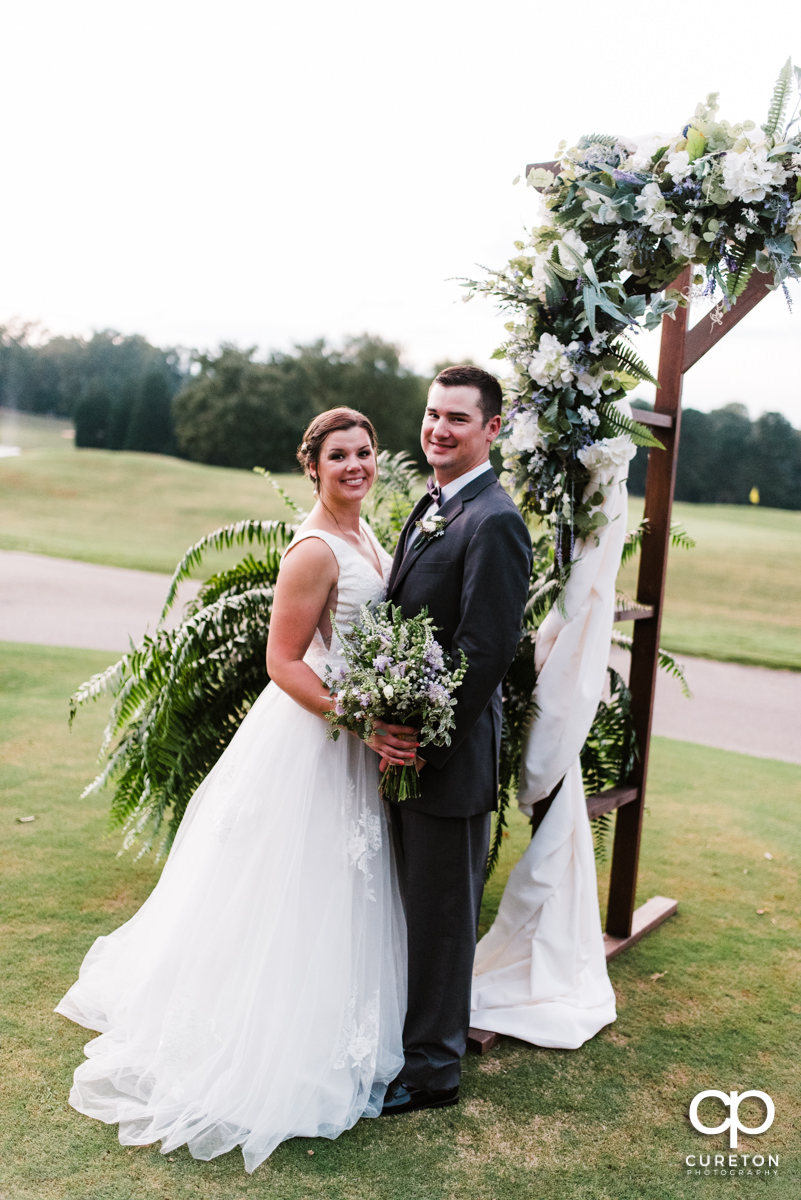 Groom and bride standing near a floral arch at their golf course wedding.