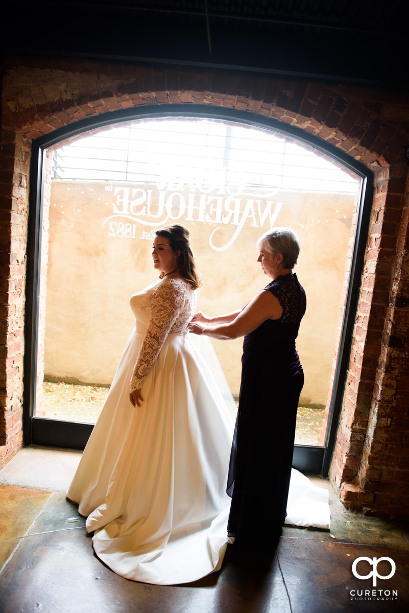 Bride's mother helping her into her dress in front of a window at The Old Cigar Warehouse.