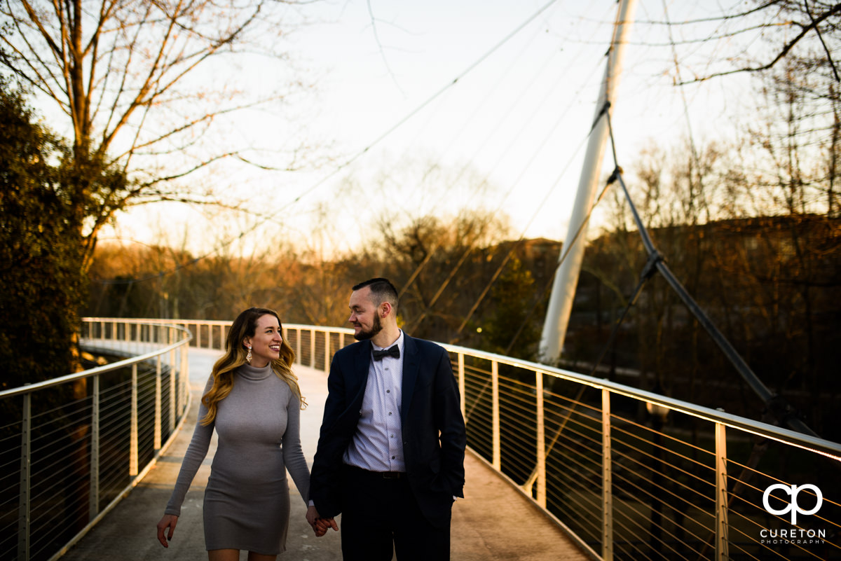 Future bride and groom walking across the Liberty Bridge in Falls Park at golden hour.