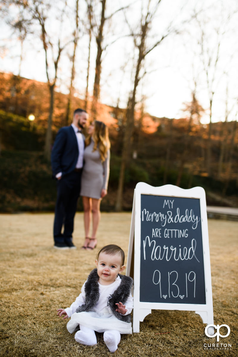 Engaged couple in the background as their baby daughter sits near a sign about their wedding for a save the date photo.