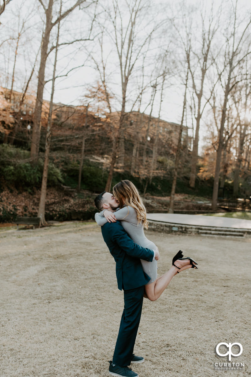 Man lifting his fiancee in the air as she kisses him.