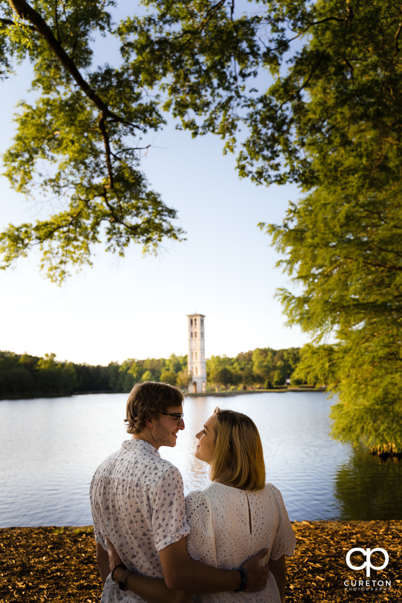 Engaged Furman students looking at each other by the lake with the Bell Tower in the background during their college graduation and engagement session at Furman University.