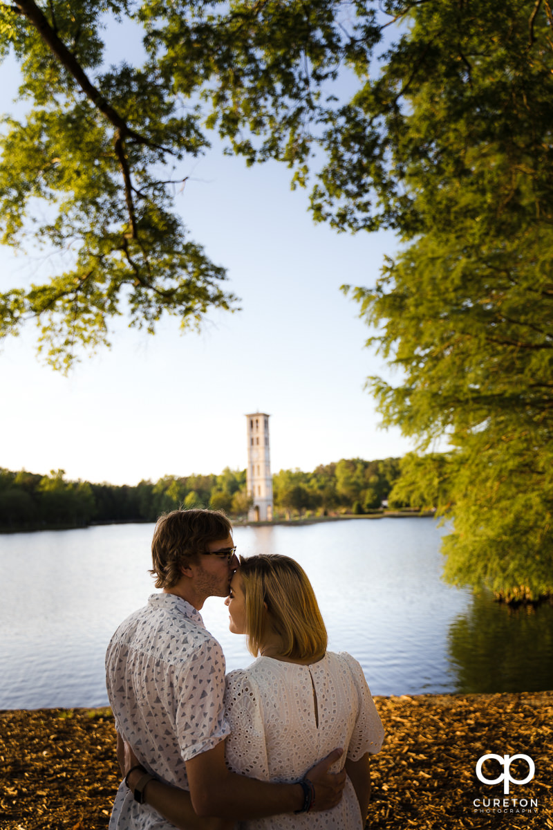 Engaged Furman students kissing each other by the lake with the Bell Tower in the background during their college graduation and engagement session at Furman University.