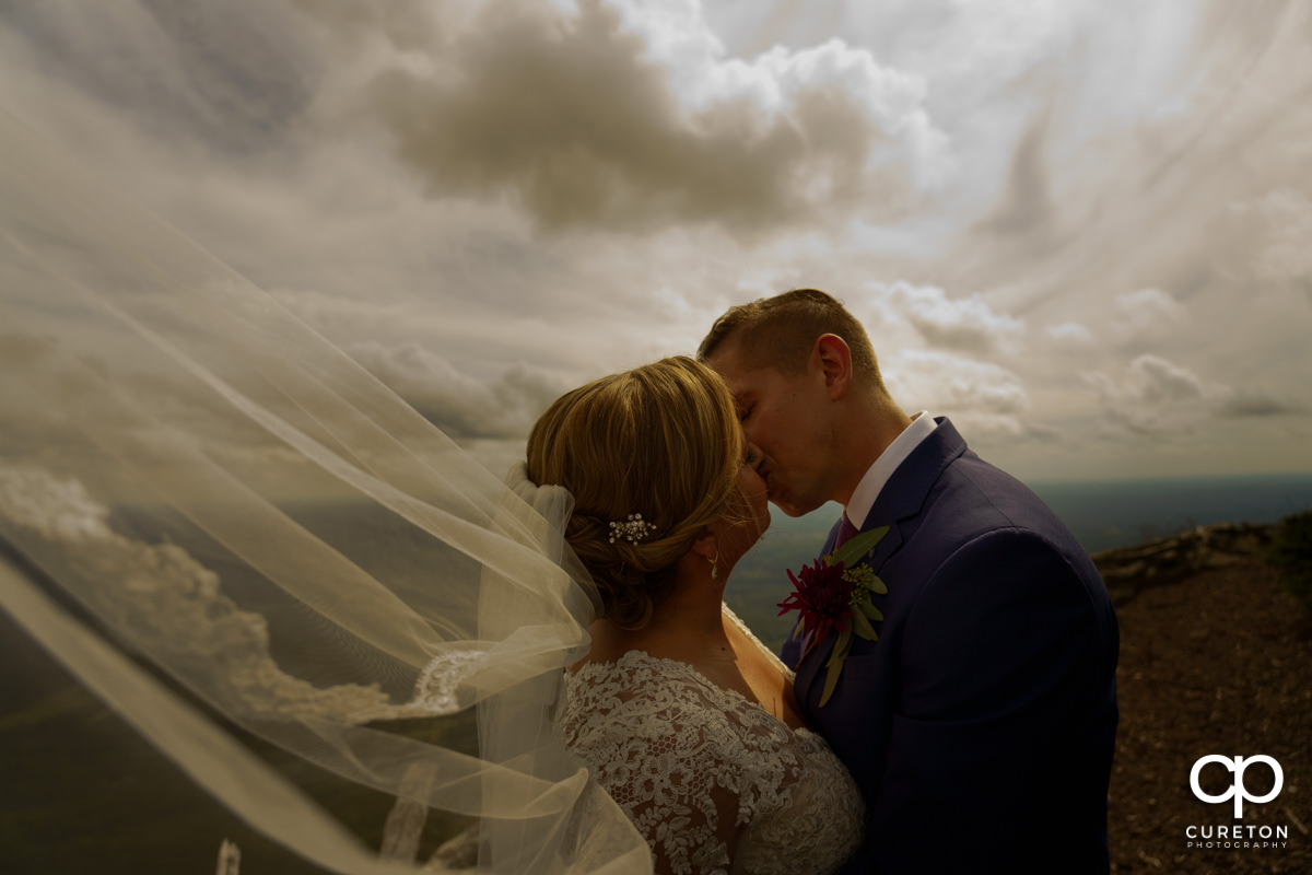 Bride and groom kissing on top of Glassy Mountain as her veil blows in the wind after their wedding at The Cliffs at Glassy.