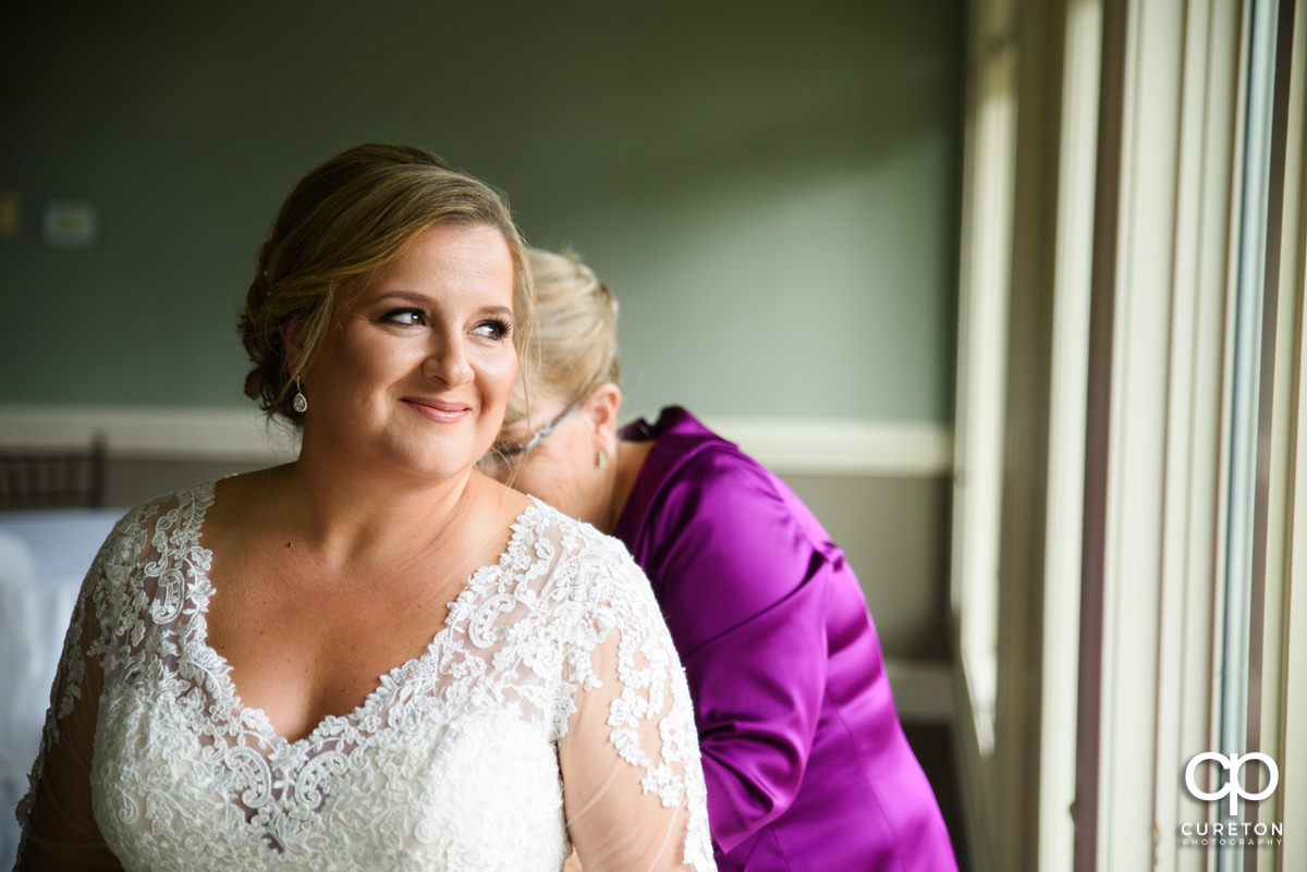 Bride looking out the window as her mother zips her dress.