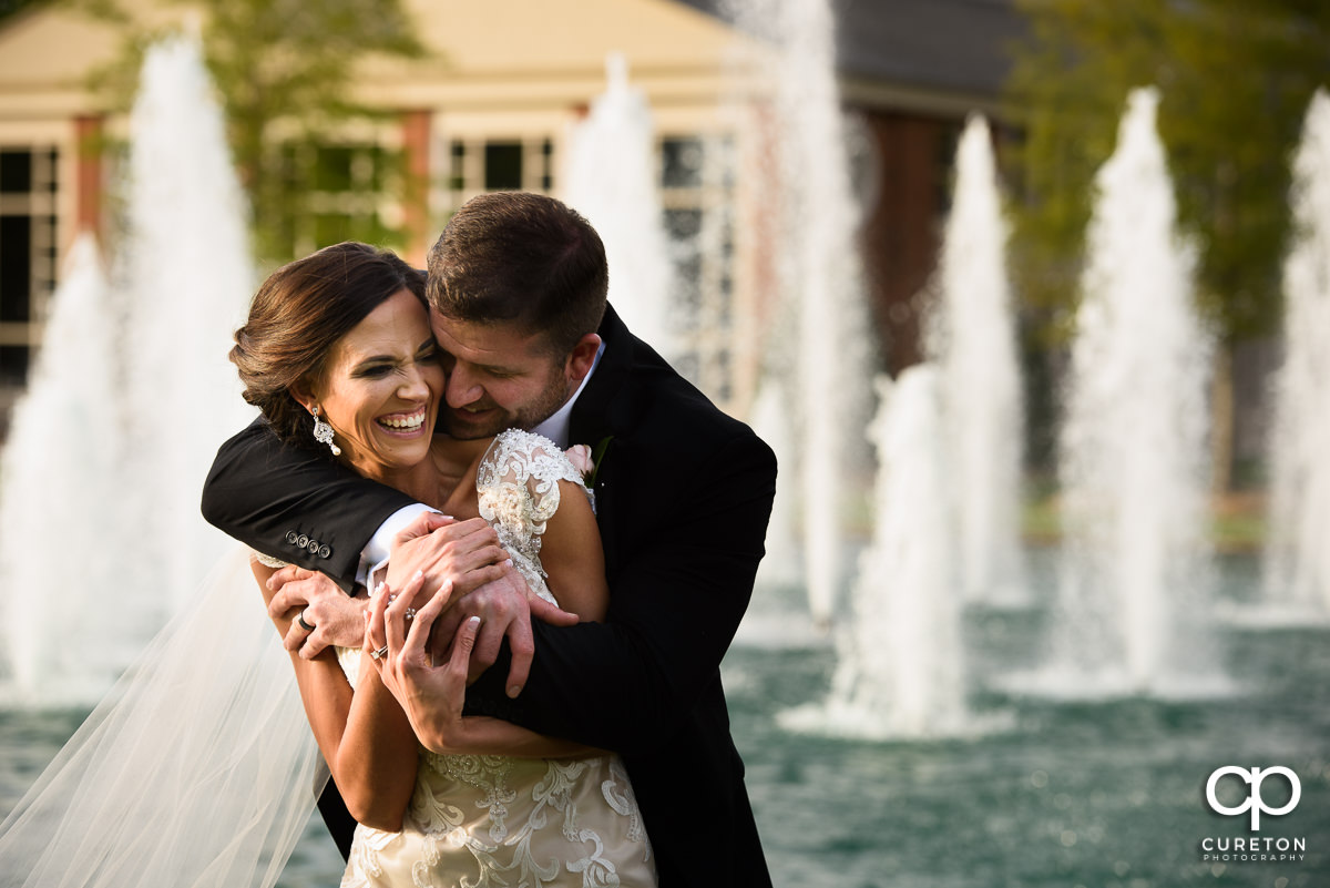 Bride smiling as her groom hugs her from behind in front of a row of fountains.