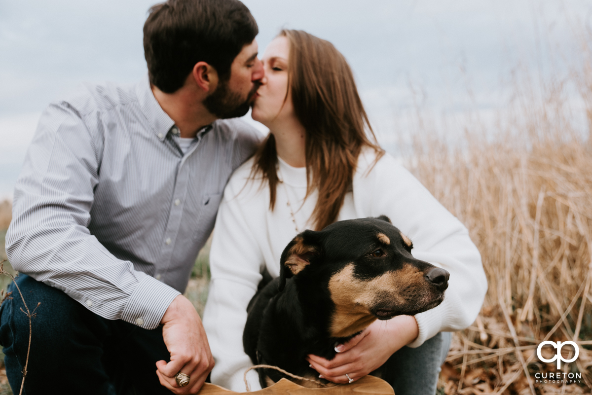 Man kissing his fiancee as their dog looks on.