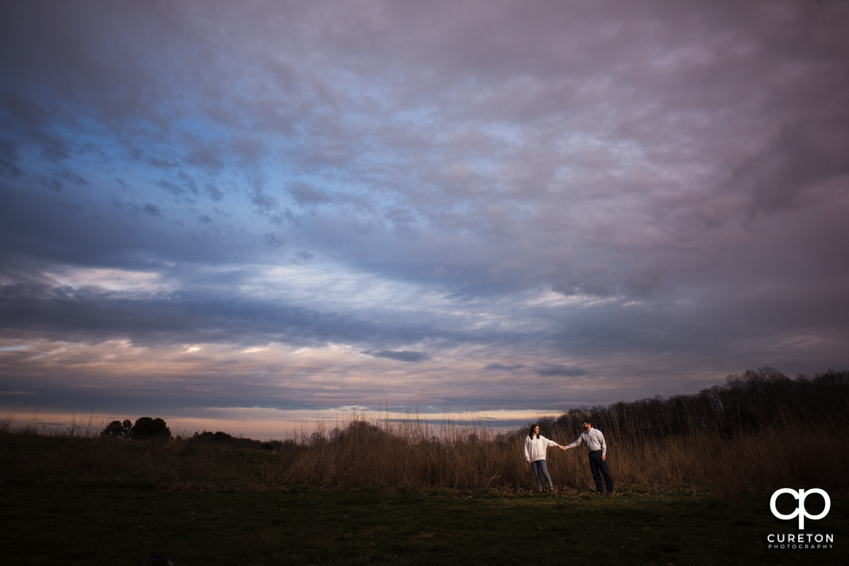 Woman leading her fiancee in a field underneath a purple sky at dusk during their engagement session.