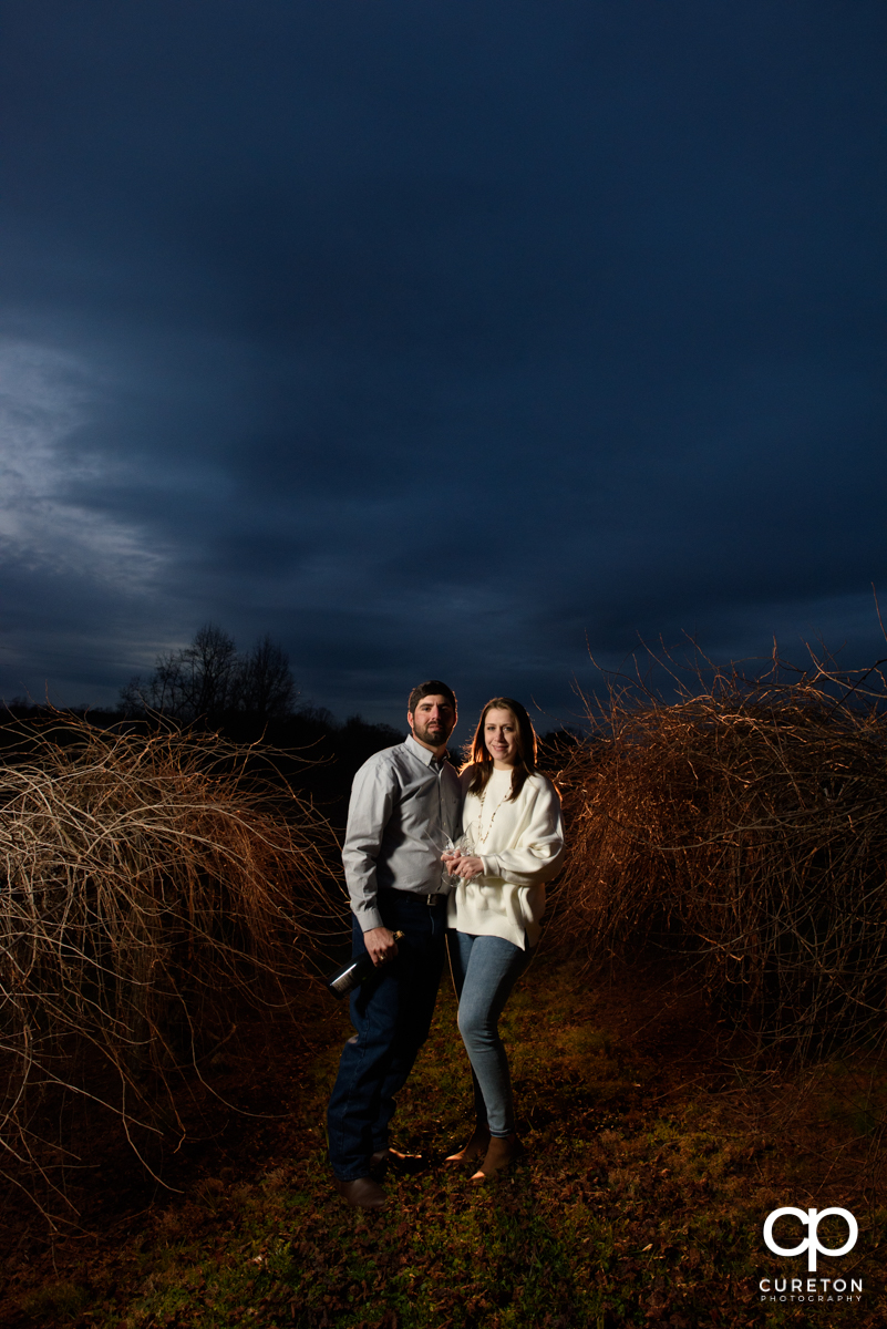 Engaged couple standing in a vineyard at sunset.