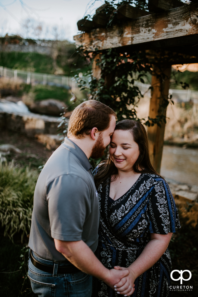 Future bride and groom snuggling near the Reedy RIver during a Greenville,SC park engagement session.