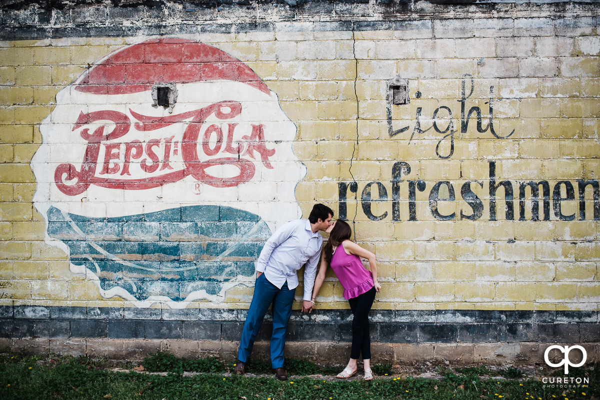 Future husband and wife kissing in front of a vintage pepsi cola sign during their engagement session in downtown Greer,SC.