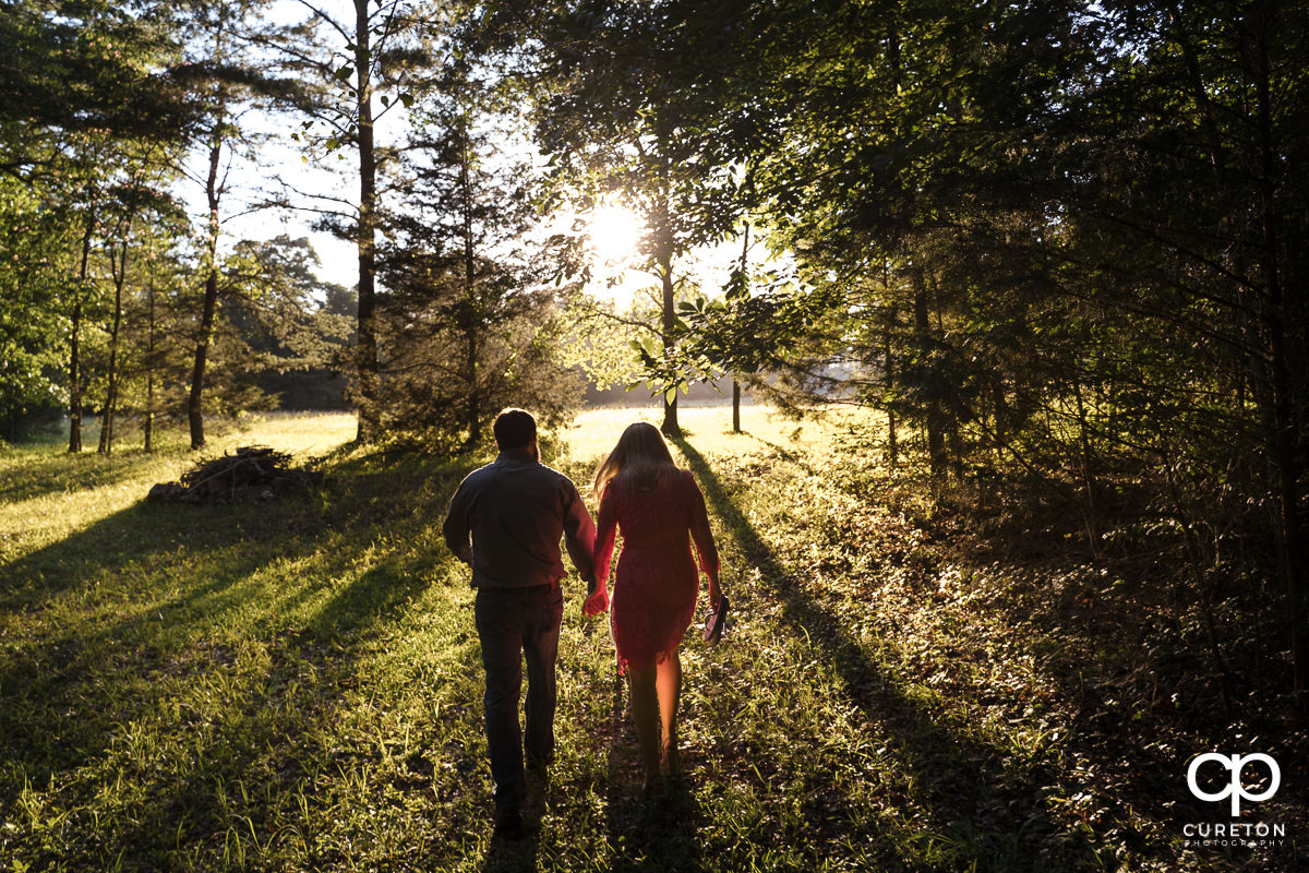 Future bride and groom walking though the woods into the sunlight during a summer engagement session in Greer,SC.