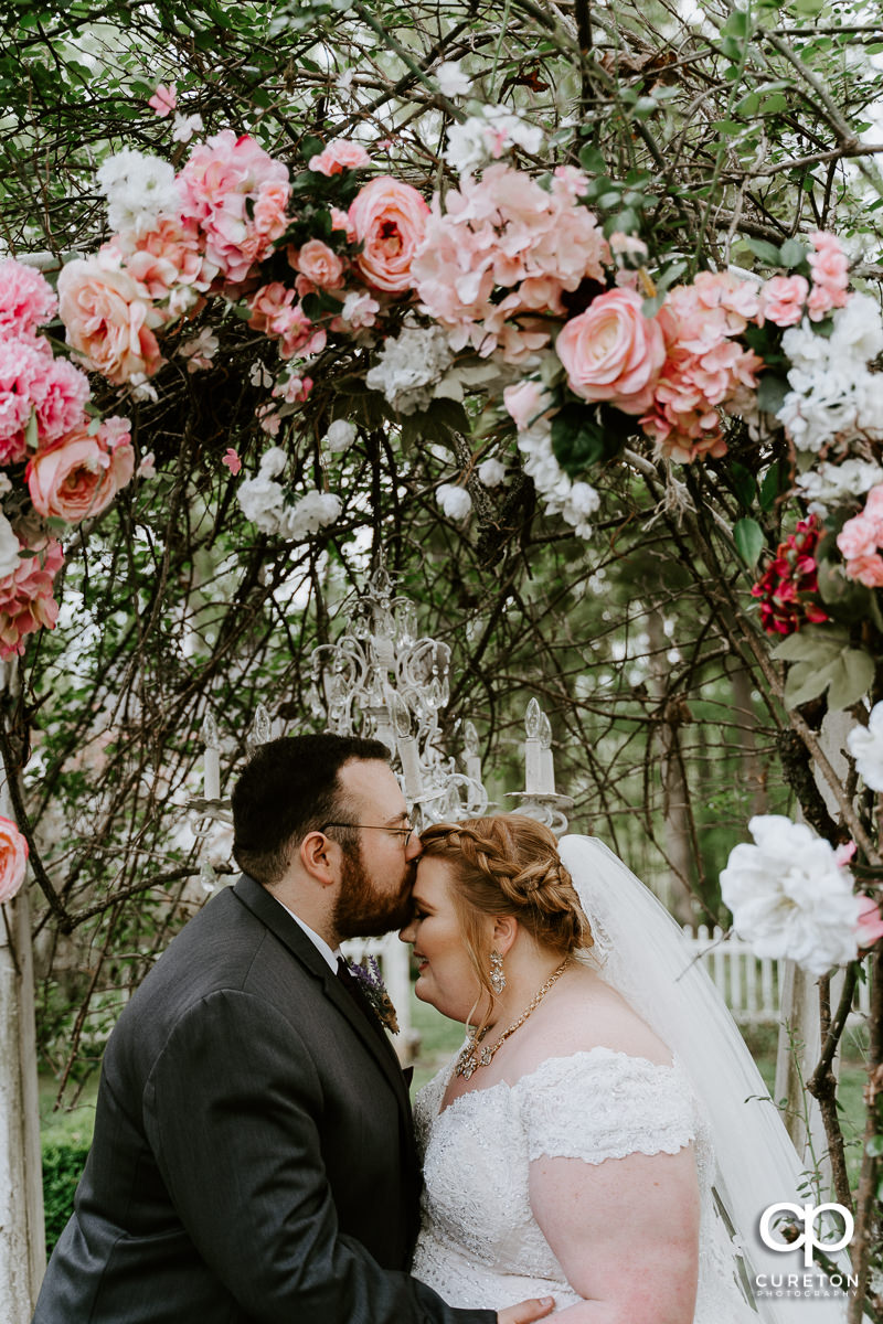 Groom kissing his bride on the forehead underneath a floral arch after their Grove at Pennington wedding in Greer,SC.