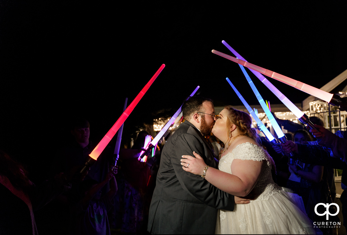 Bride and groom kissing with the wedding party holding light sabers over them.