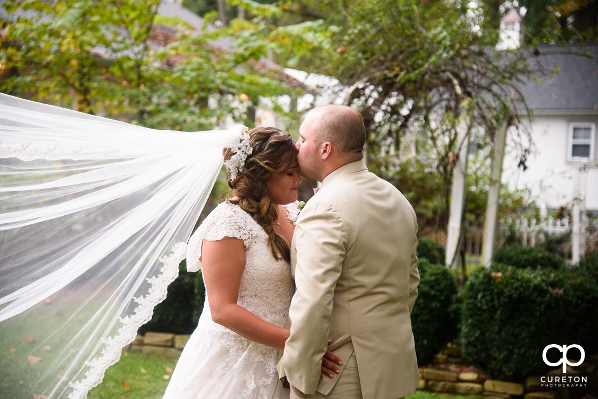 Groom kissing his bride on the forehead with her veil blowing in the wind.