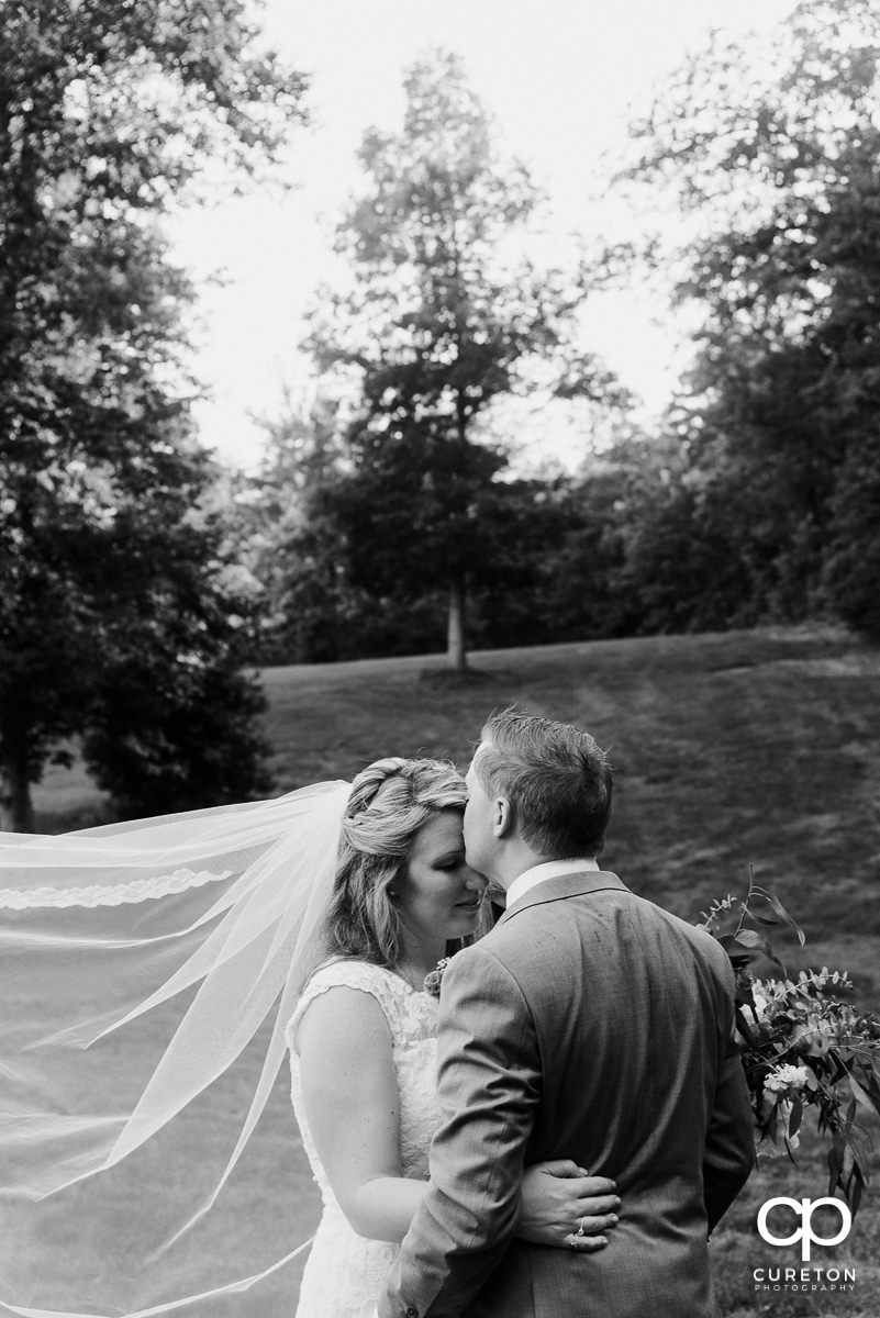 Groom kissing his bride on the forehead as her veil blows in the wind.