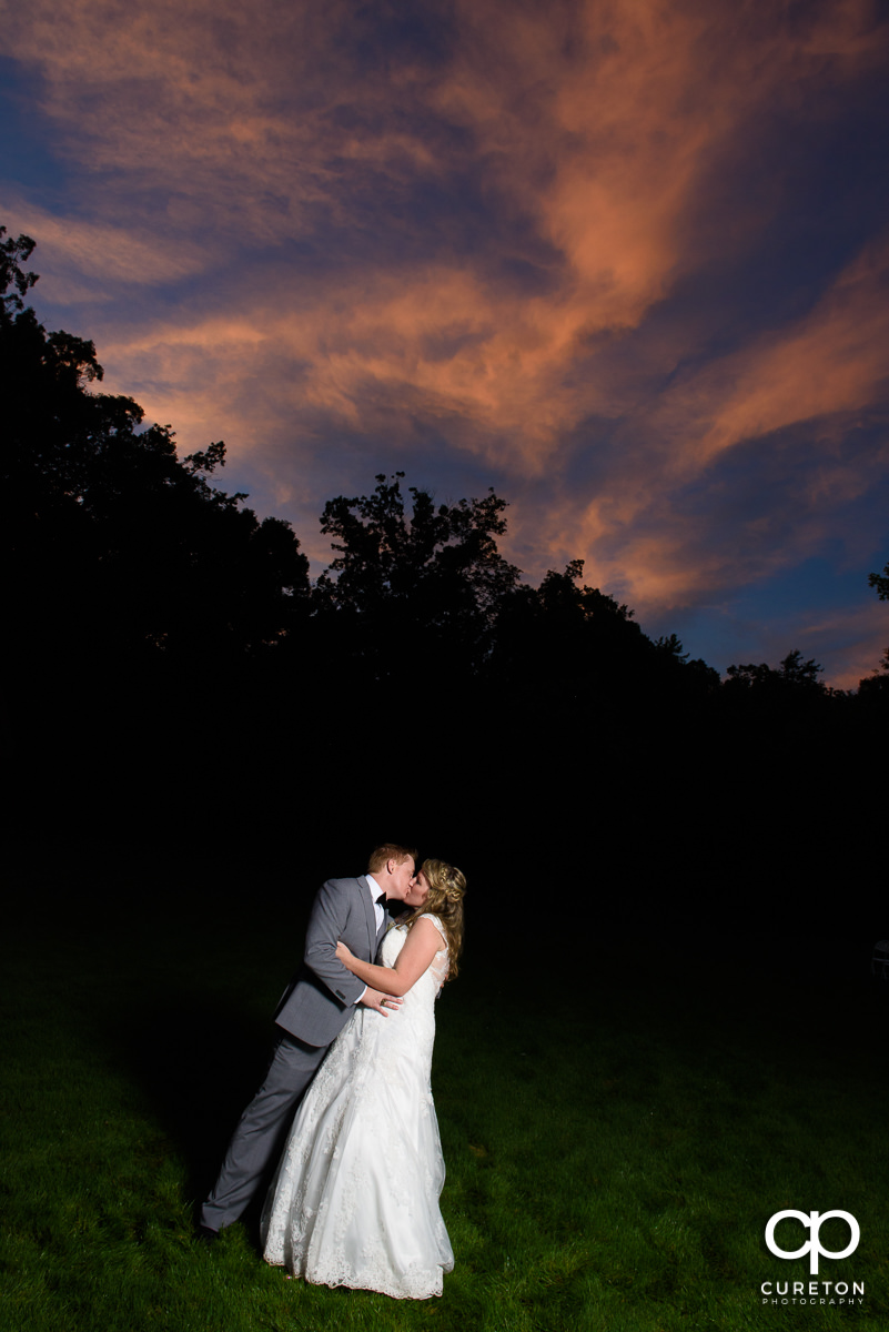 Bride and groom kissing under a sunset sky.