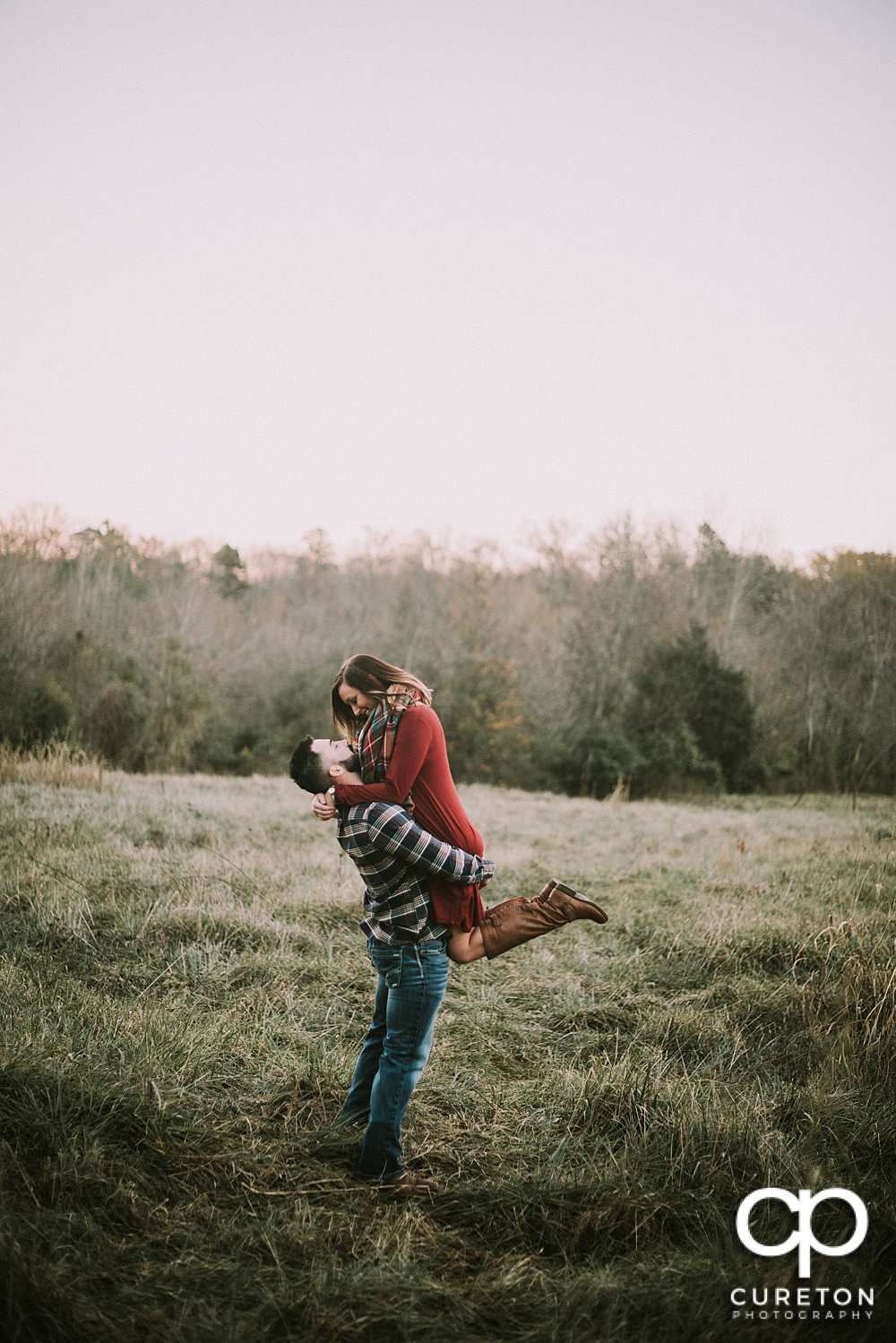 Man lifting his fiance in a field during their Lake Conestee Nature Park engagement session.