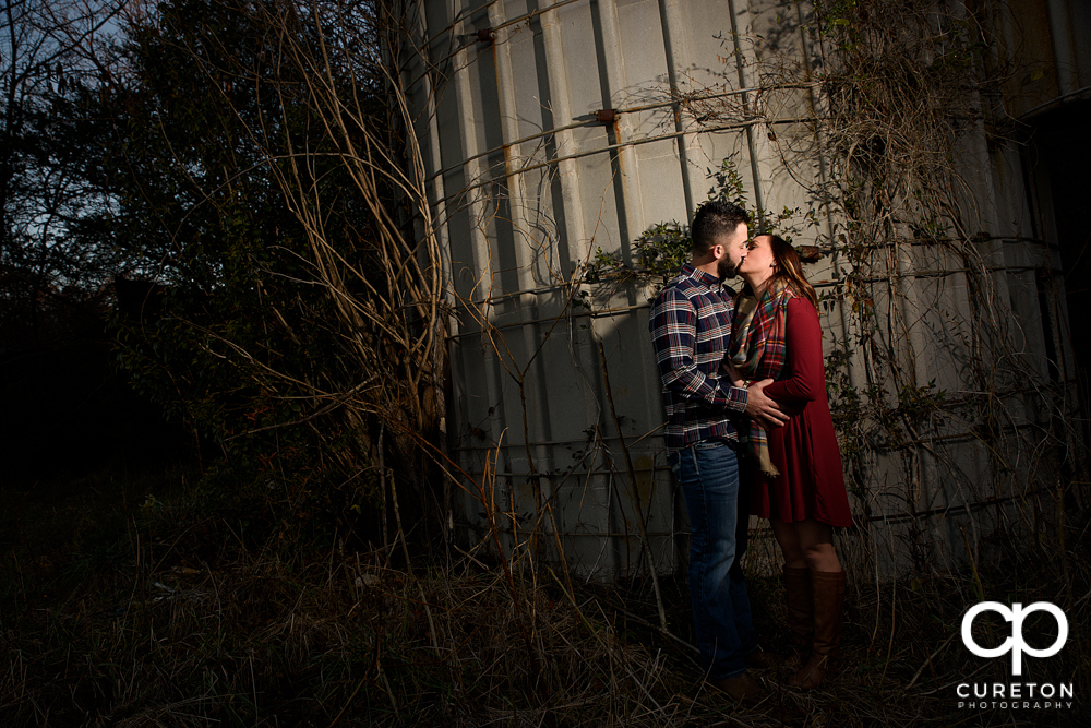 Engaged couple kissing while leaning on a corn silo.