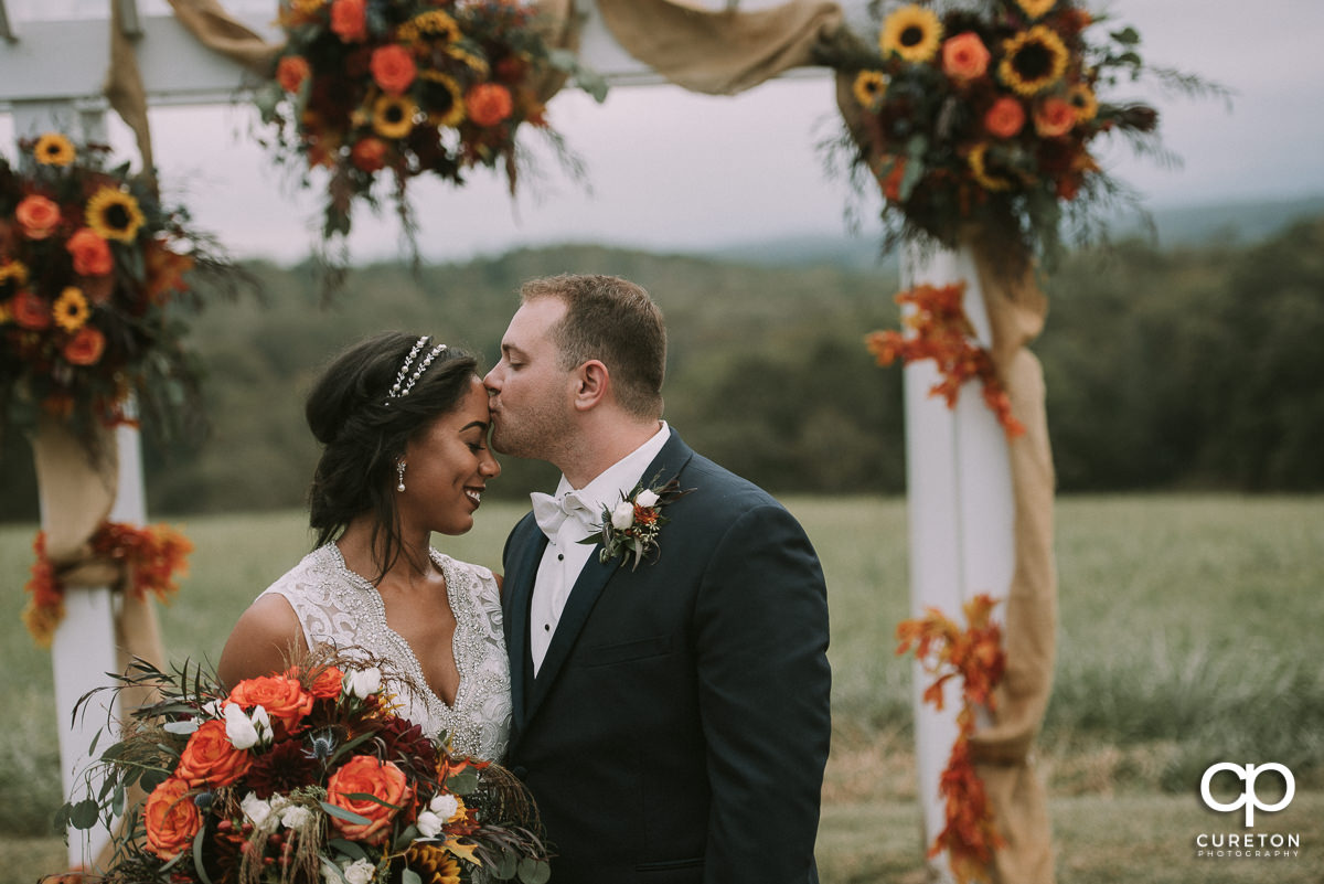 Groom kissing his bride on the forehead with beautiful fall flowers in the background at their Lindsey Plantation wedding.