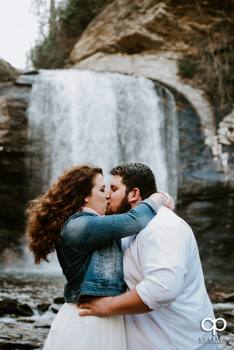 Engaged couple kissing in front of a waterfall.