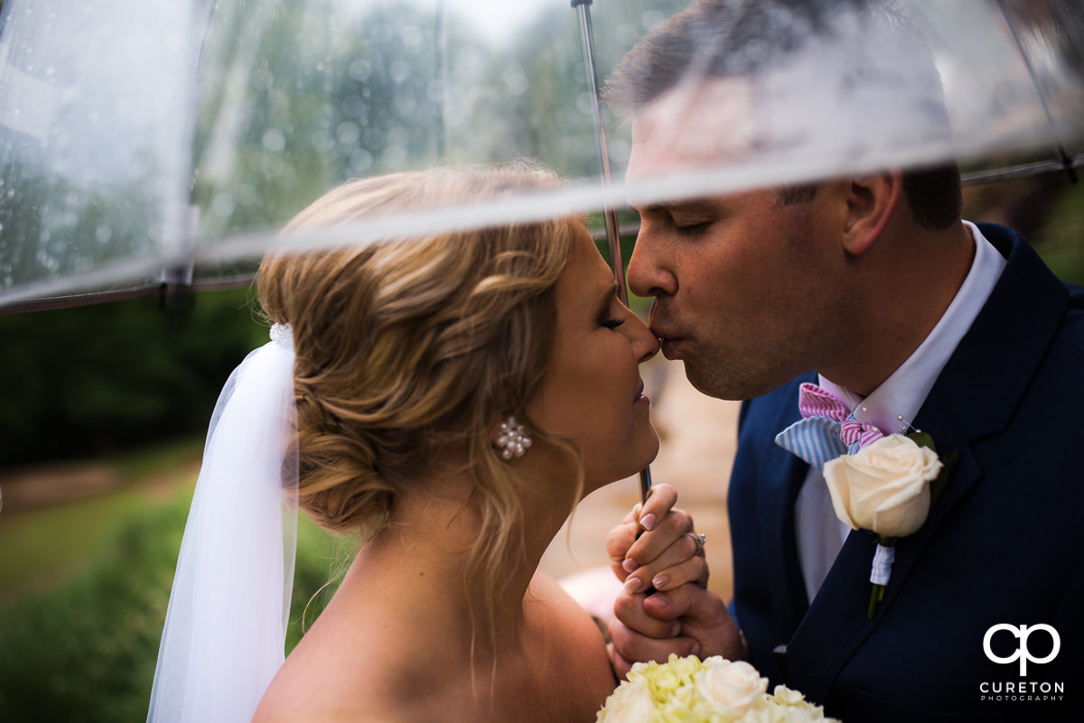 Groom kissing his bride on the noes underneath an umbrella in the rain before their Mary's at Falls Cottage wedding in downtown Greenville,SC.