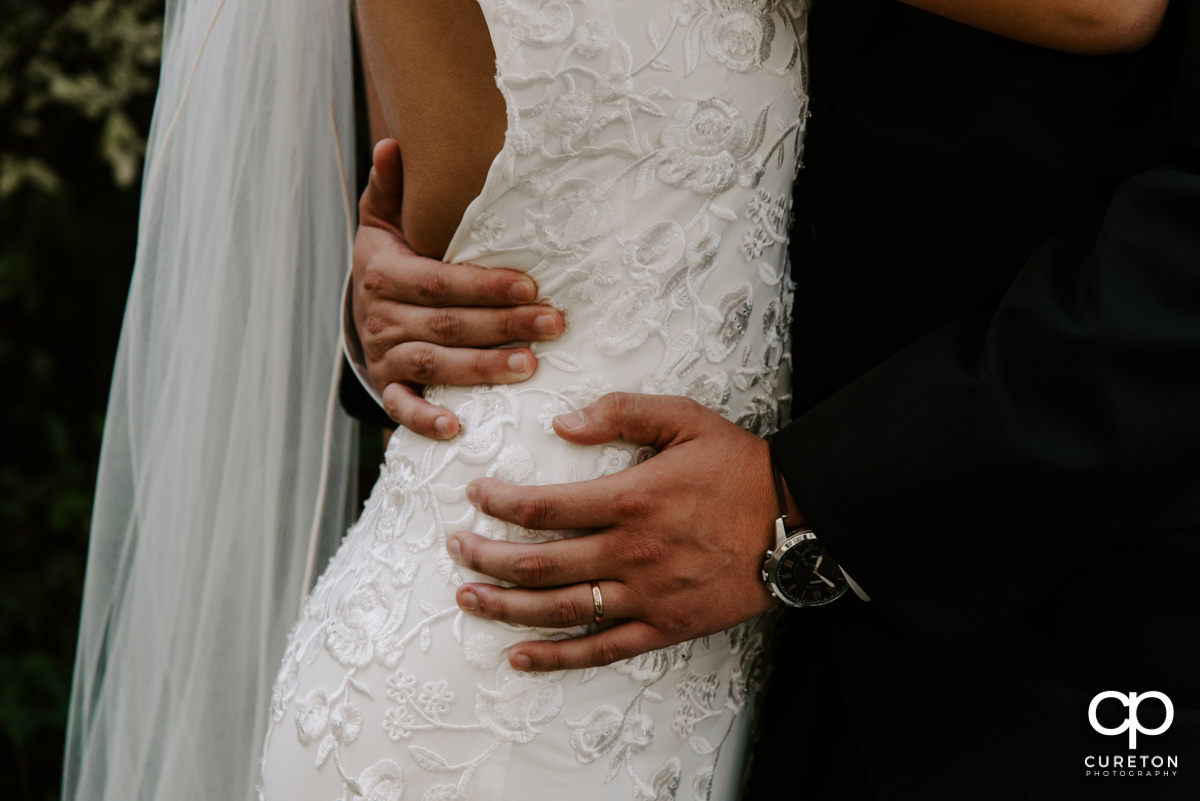 Closeup of groom's hands and ring as he holds his wife's waist.