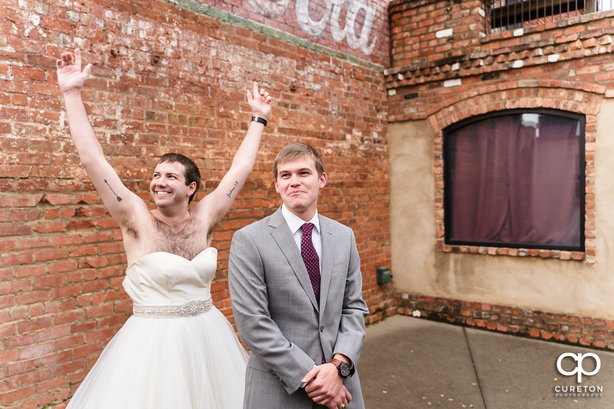 Groomsmen in a wedding dress showing up to the first look to trick the groom at The Old Cigar Warehouse as the bride looks on.