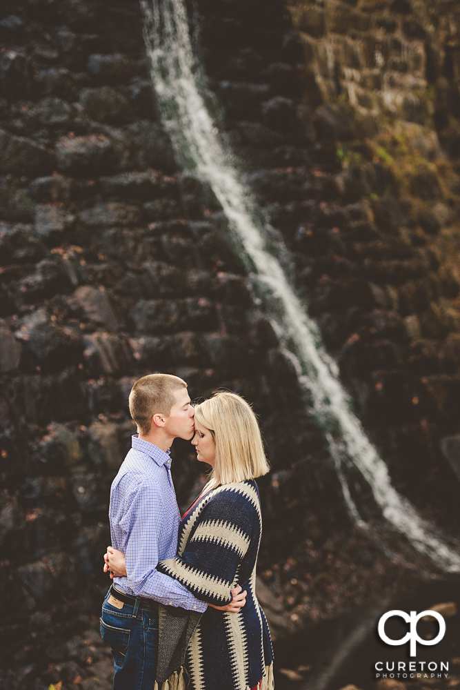 Man kissing his fiancée on the forehead in front of the dam at Paris Mountain State Park during their engagement session.