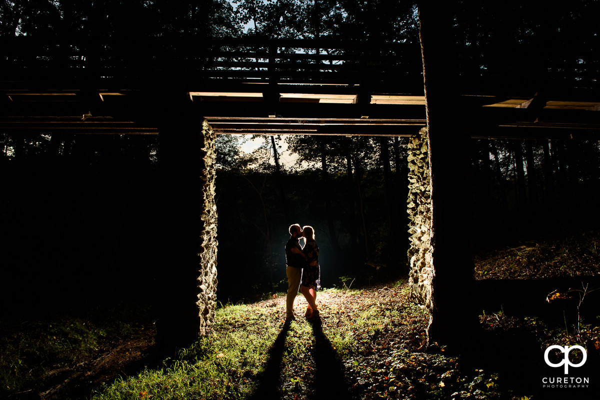 Backlit shot of a bride and groom underneath a walking bridge in the woods at an engagement session at Paris Mountain State Park in Greenville,SC.